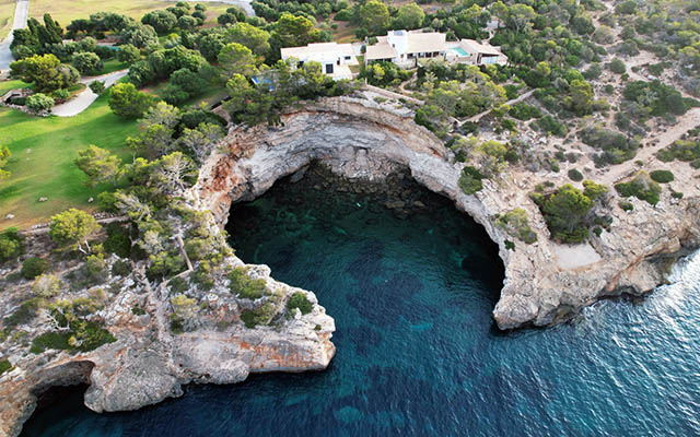 Luftaufnahme einer felsigen Küste mit einer großen Höhle, klarem blauem Wasser und einem weißen Gebäude umgeben von Bäumen.