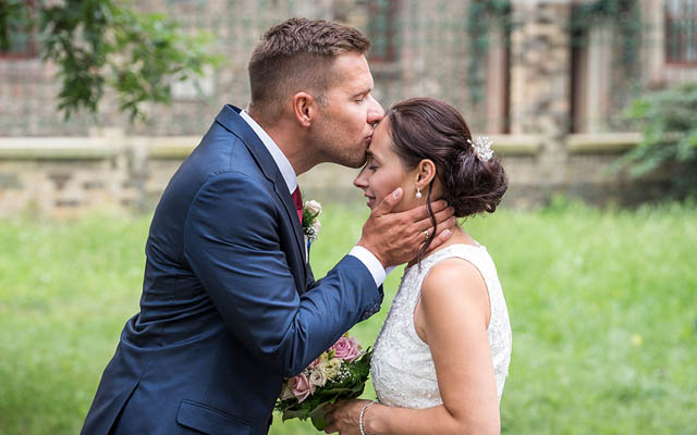 Bräutigam im dunkelblauen Anzug küsst Braut in weißem Spitzenkleid, Braut hält Blumenstrauß, im Garten.