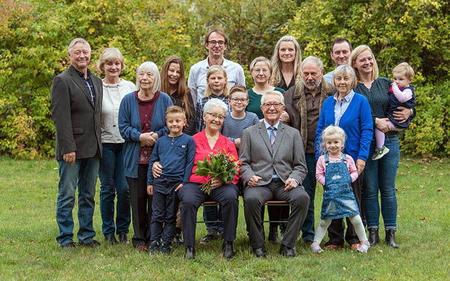 Gruppe von Erwachsenen und Kindern im Garten, zwei ältere Personen sitzen mit Blumenstrauß, umgeben von stehenden Familienmitgliedern.