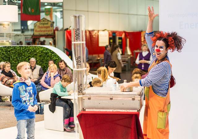 Clown mit rotem Haar und orangefarbenem Overall steht an Tisch mit transparenten Messzylindern, mehrere Kinder und Erwachsene sitzen und stehen im Hintergrund.