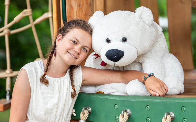 Kind mit geflochtenem Haar und weißem Kleid lehnt an grünem Klettergerüst neben großem weißem Teddybär mit roter Schleife.